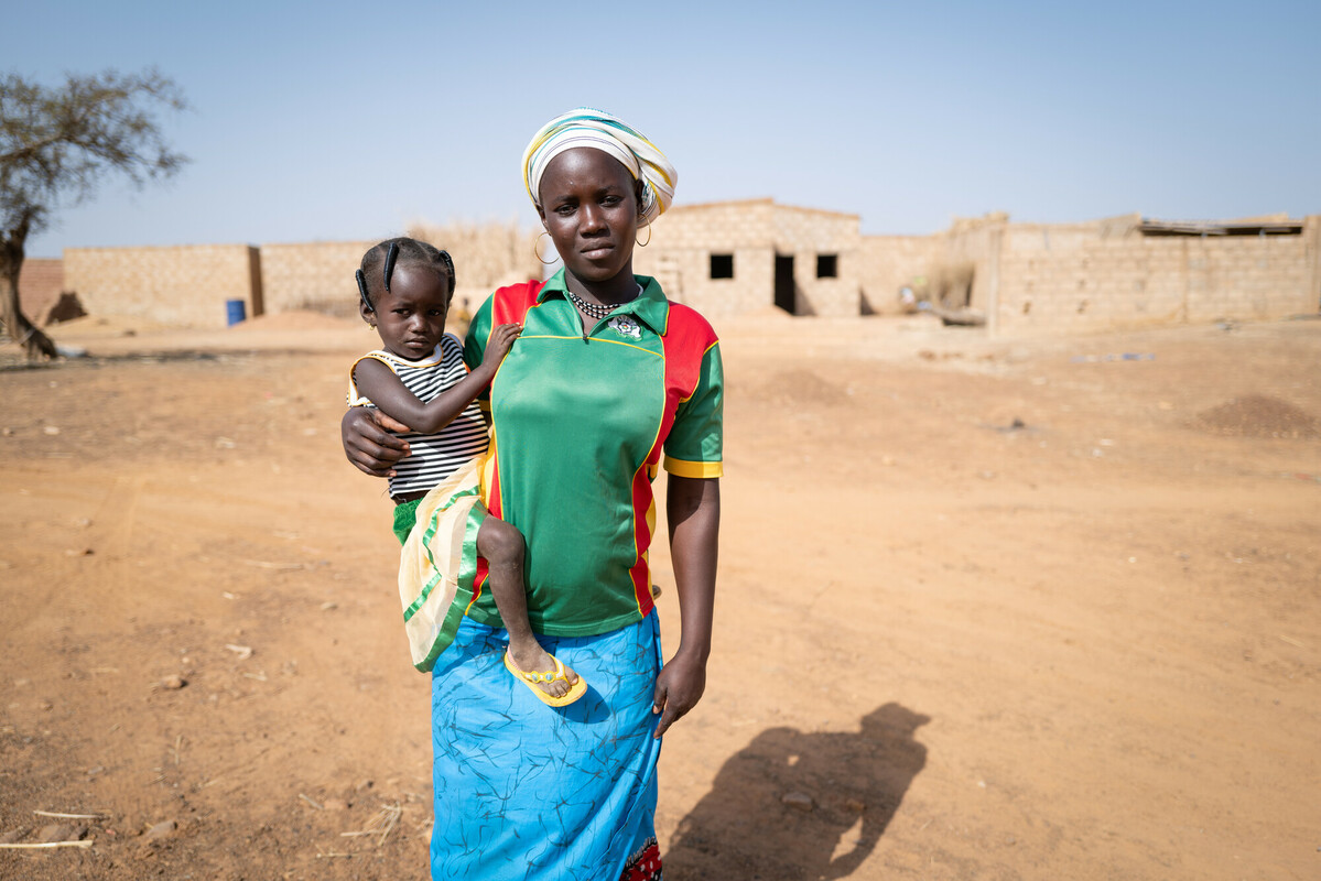 Mariam holds her daughter in her arms in the Kaya internal refugee camp in Burkina Faso. Mariam is wearing a green sports shirt and a blue skirt.