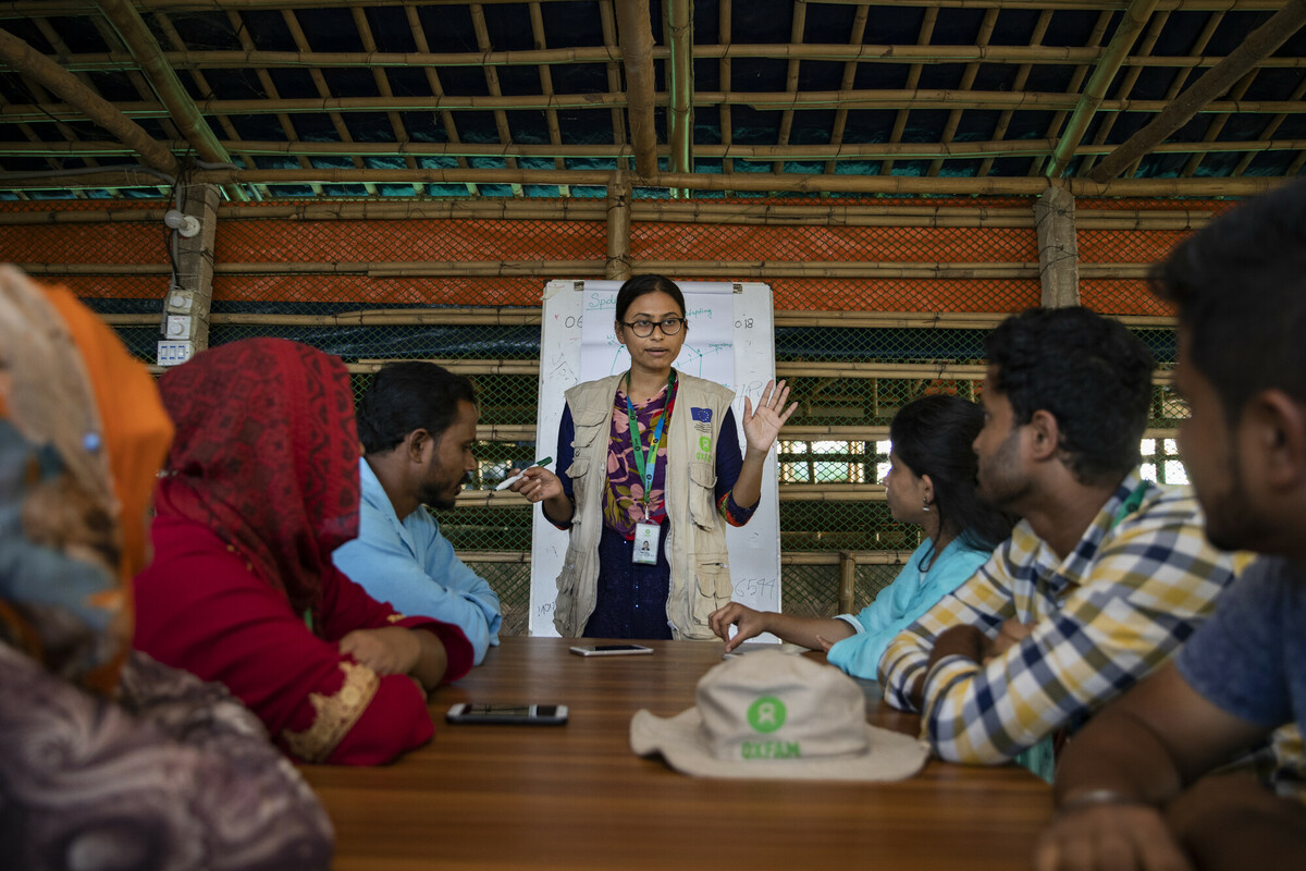 En kvinna står vid fotändan av ett bord och föreläser inför en grupp av människor som sitter runt bordet, Bangladesh.