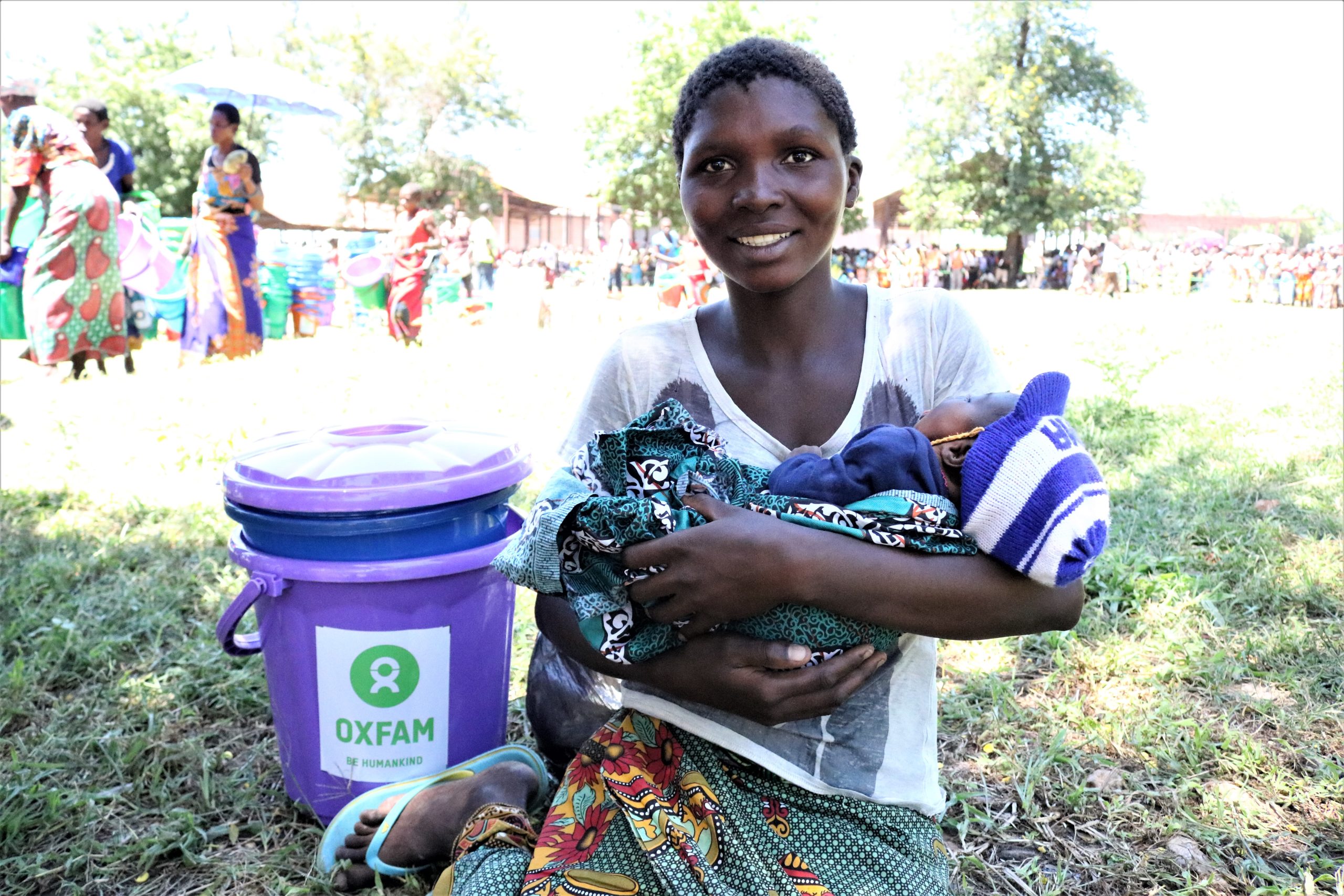 Zainabu håller sitt barn i famnen. I bakgrunden står en vattenhink från Oxfam. Bilden är tagen i Bangula lägret.